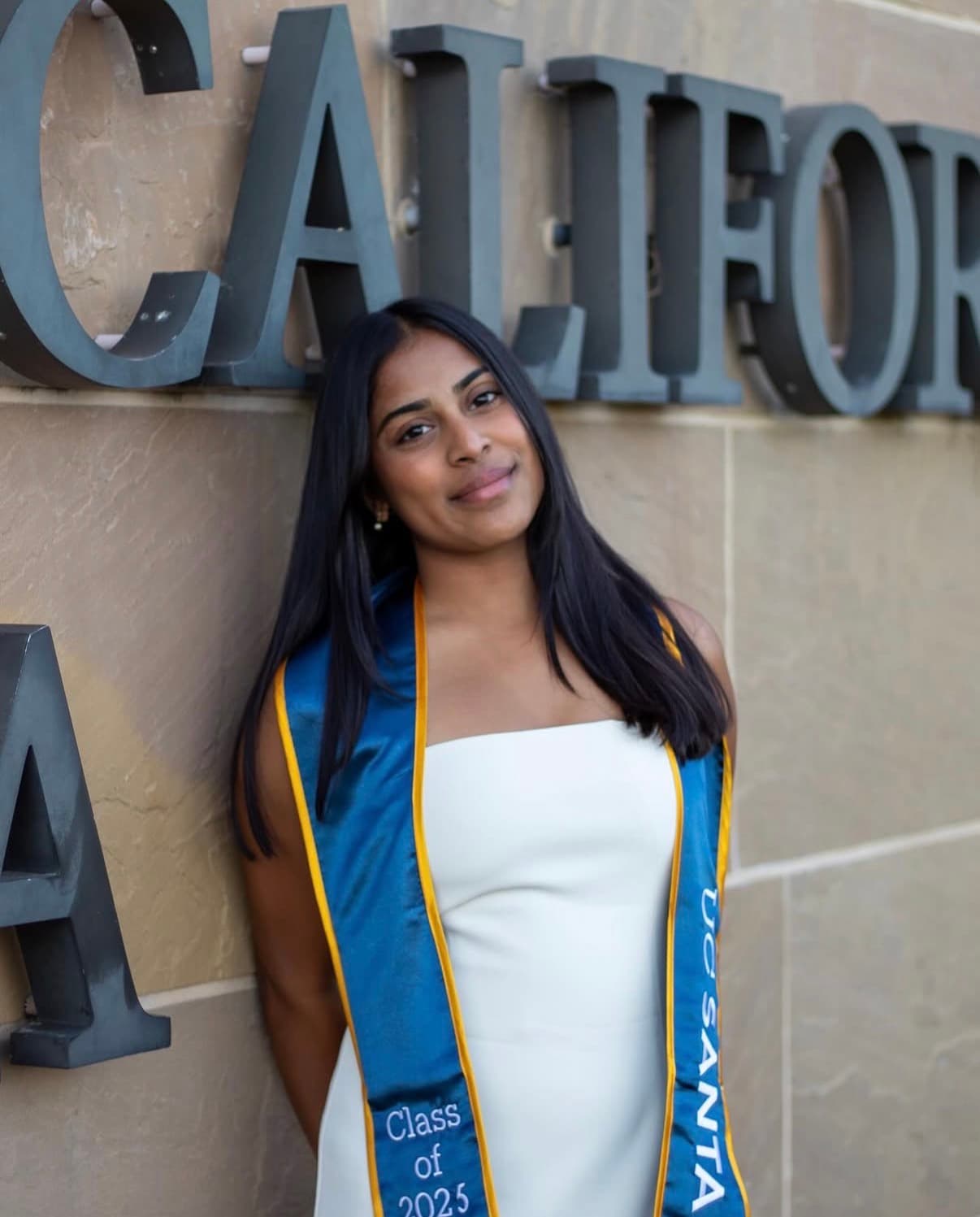 Graduate portrait close to the California wall with a soft smile and blue stole.