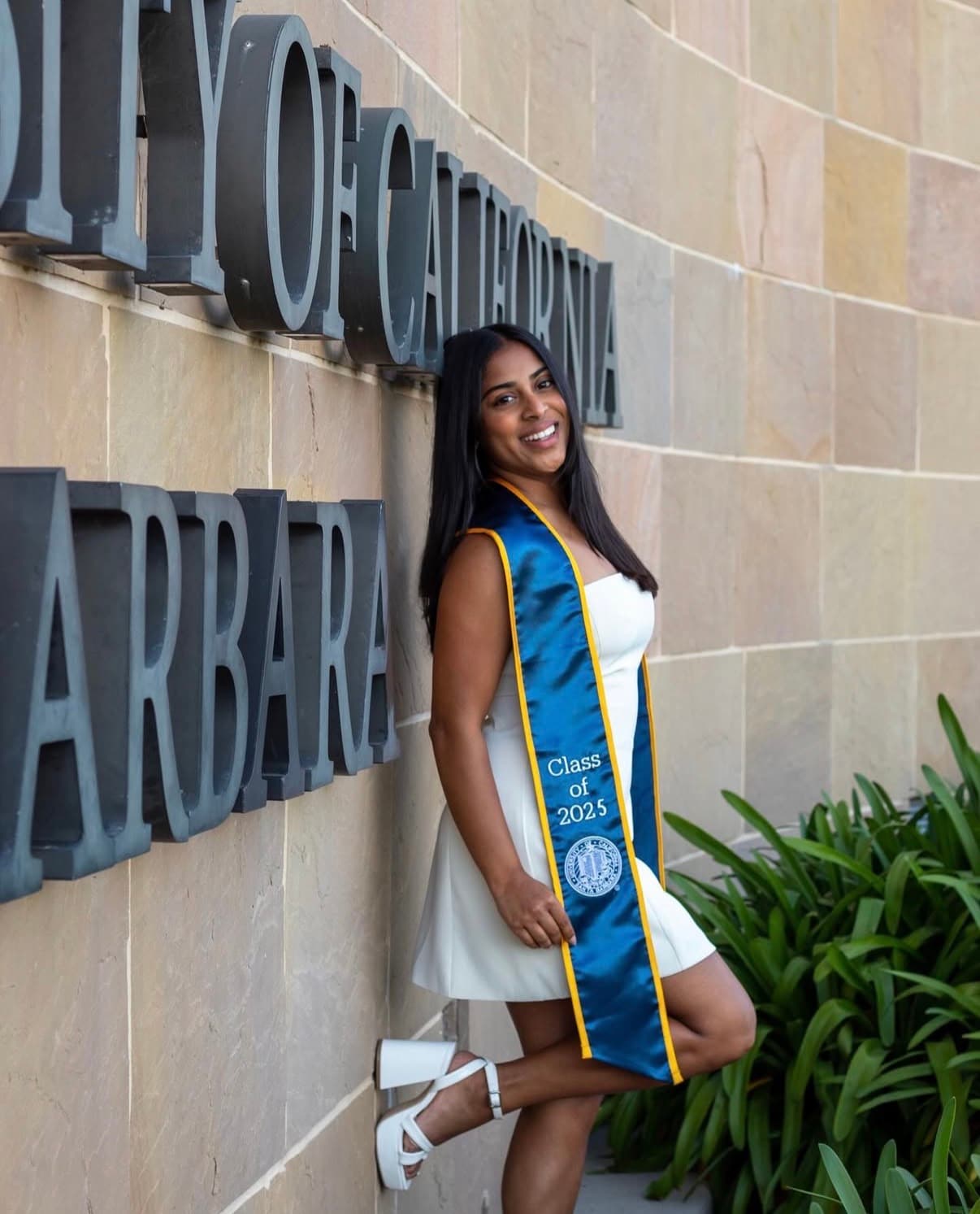 Graduate in a white dress posing full length beside California campus lettering.