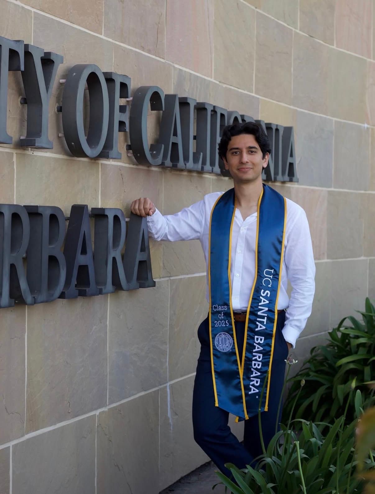 Graduate leaning beside California signage in a tailored white shirt and stole.