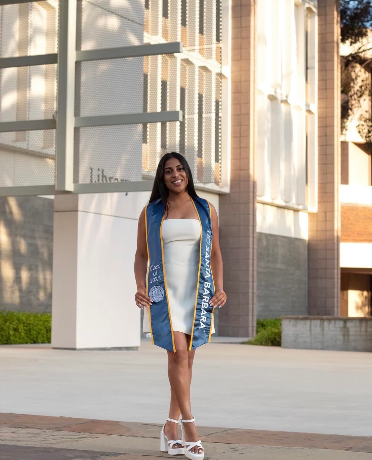 Graduate walking through a campus courtyard in evening light with her UCSB stole.