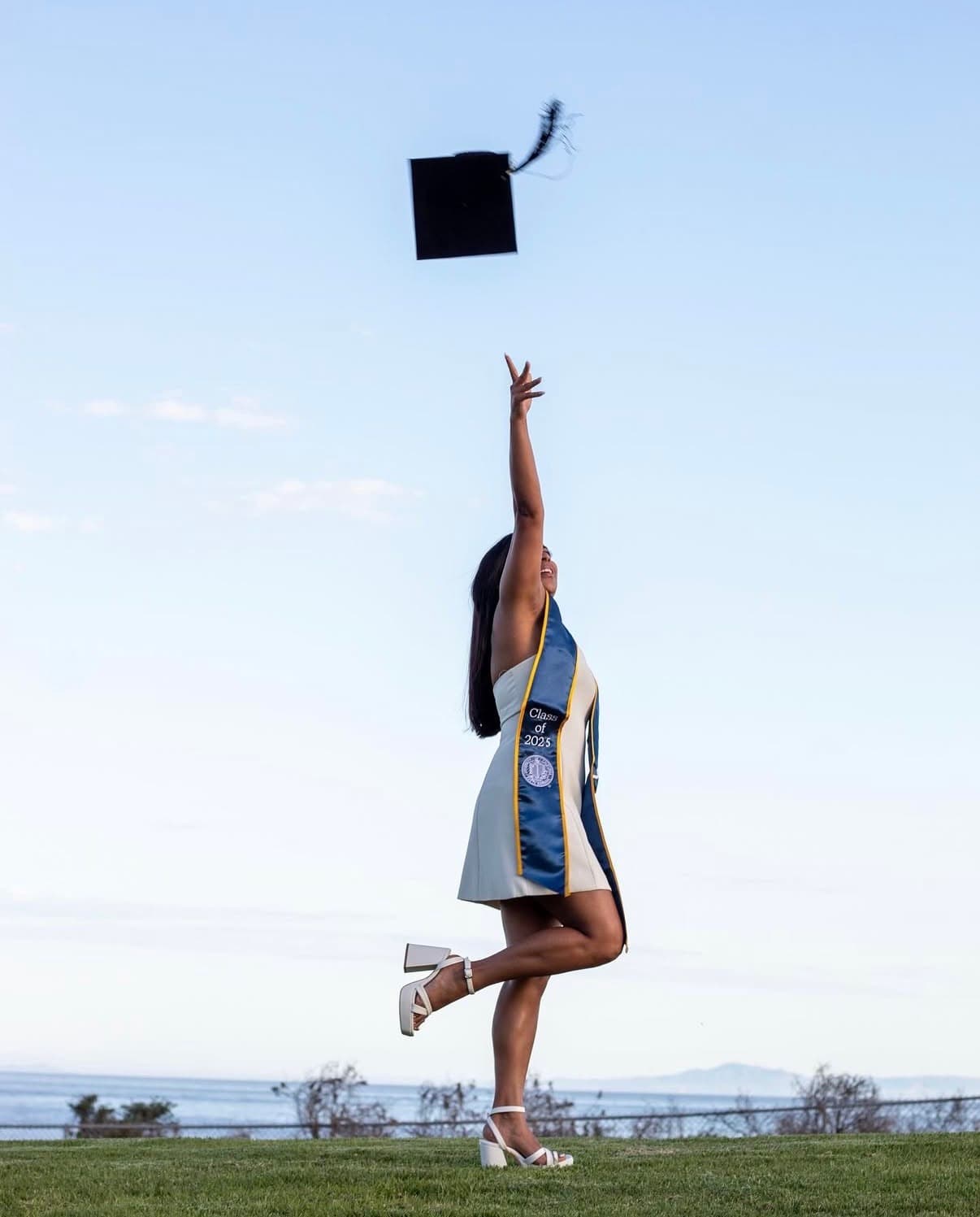 Graduate reaching for a tossed cap on a grassy bluff above the water.