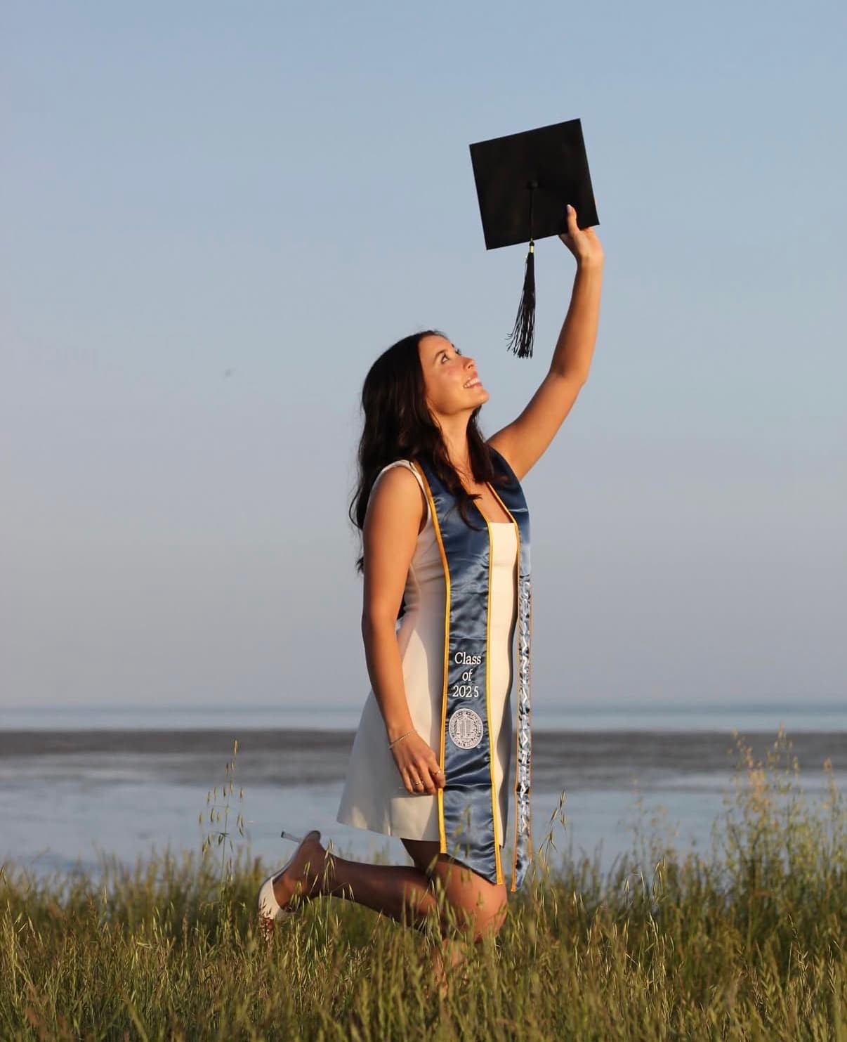 Graduate lifting her cap overhead while kneeling in shoreline grass at sunset.