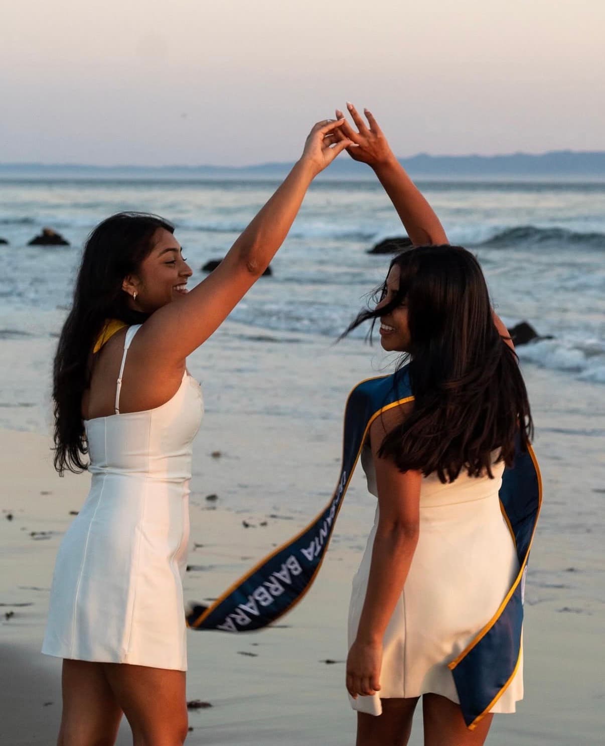 Two graduates dancing together barefoot at the shoreline during golden hour.