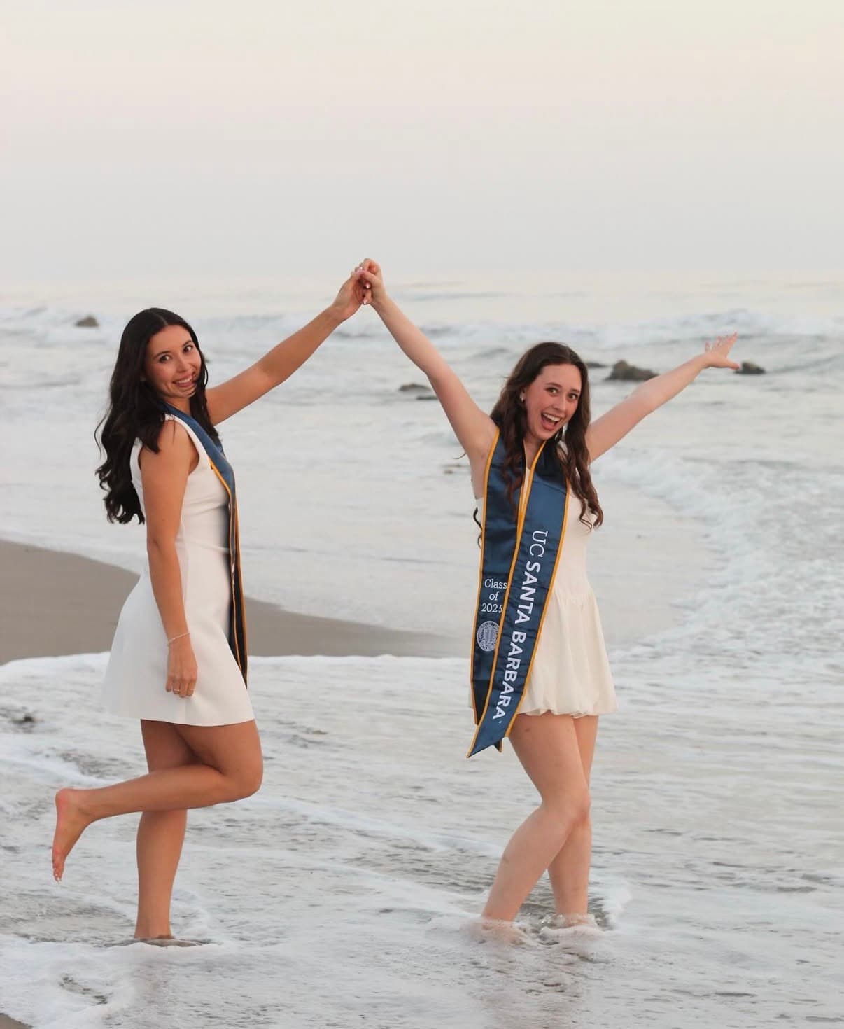 Two graduates holding hands and celebrating together in the surf at dusk.
