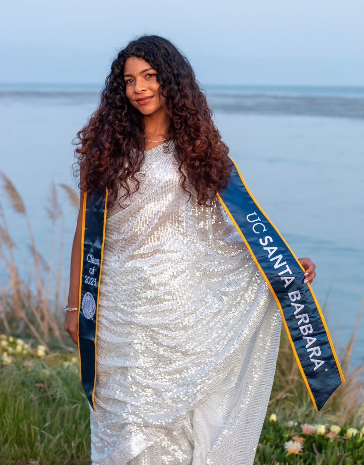 Graduate in a silver gown and UCSB stole posing along the shoreline at blue hour.