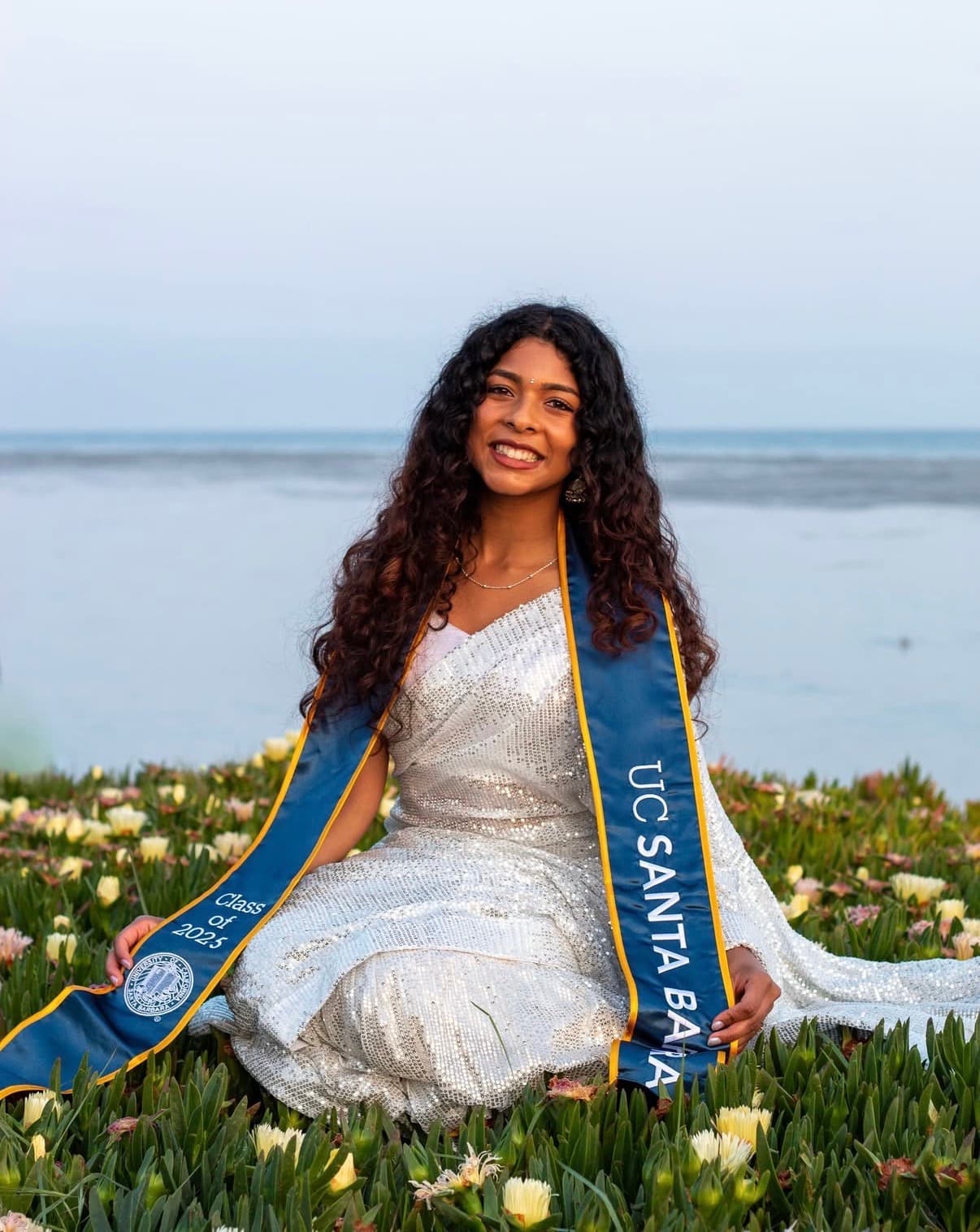 Graduate seated among coastal blooms in a silver gown with the ocean behind her.