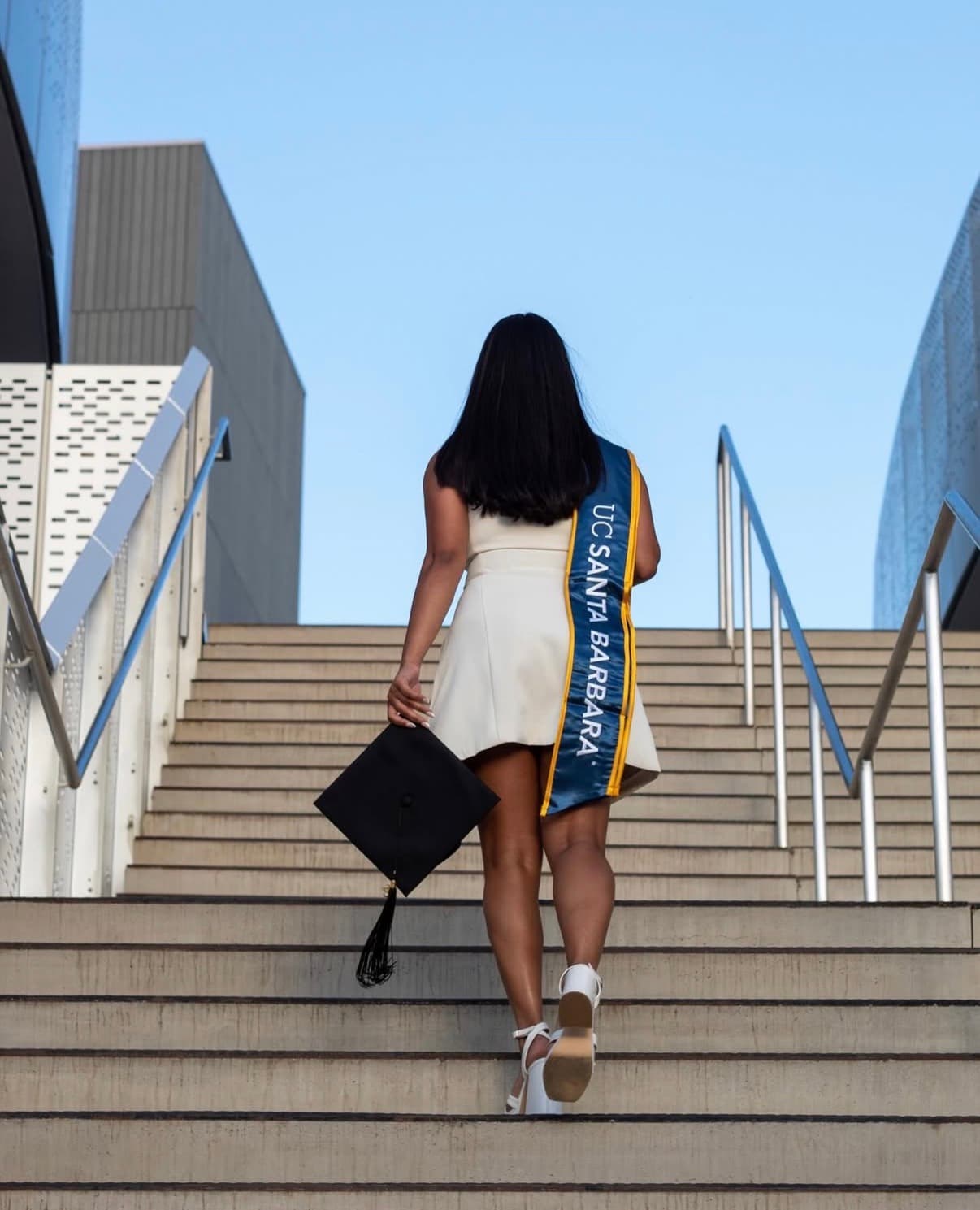 Graduate walking up campus stairs with her cap in hand and stole trailing behind.