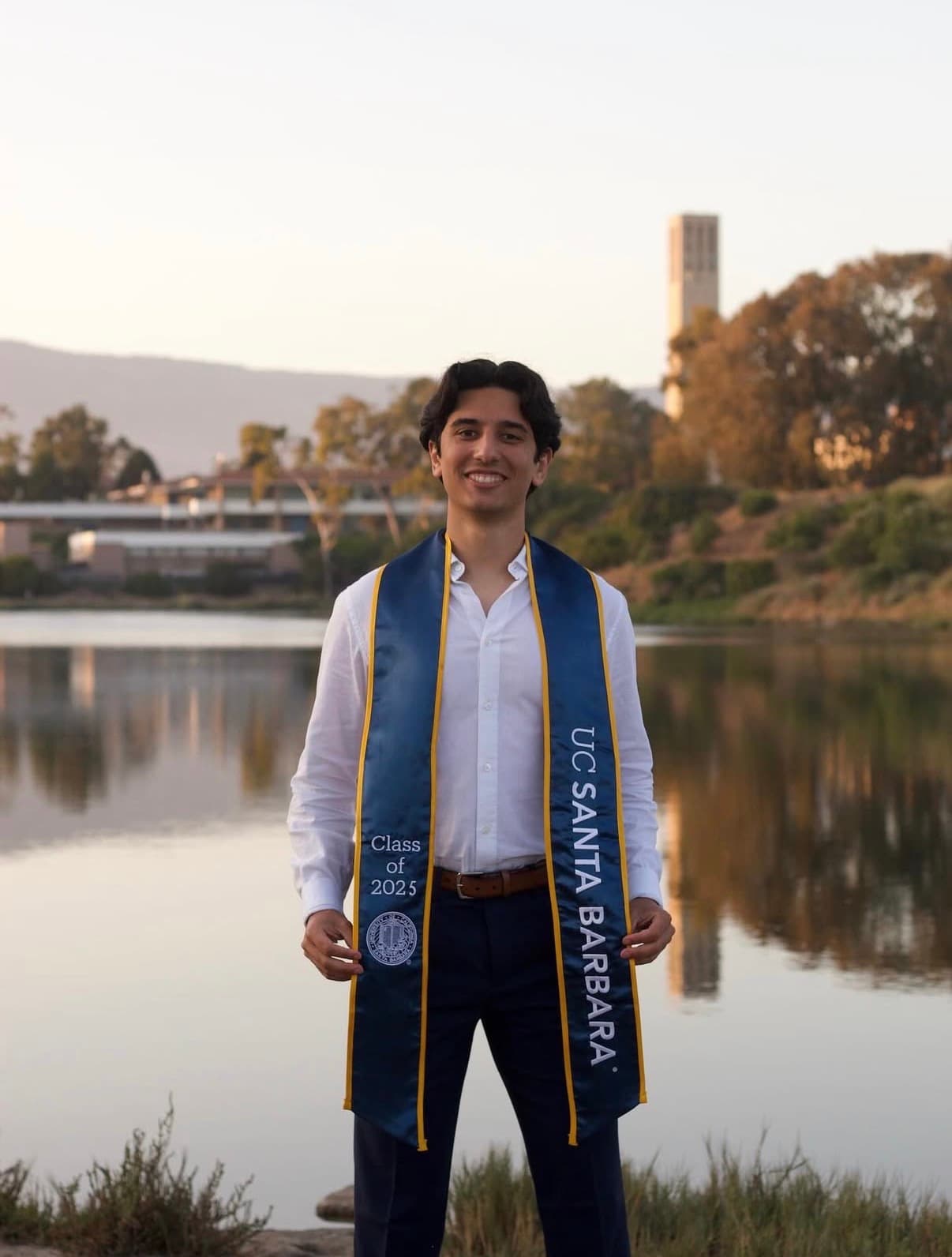Graduate in a white shirt and UCSB stole standing beside the water at sunset.