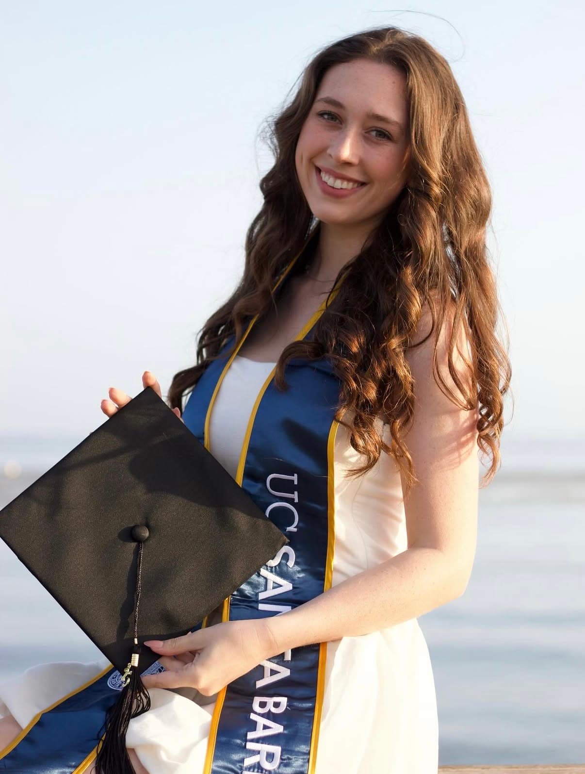 Graduate holding her cap against a pale coastal sky in soft evening light.