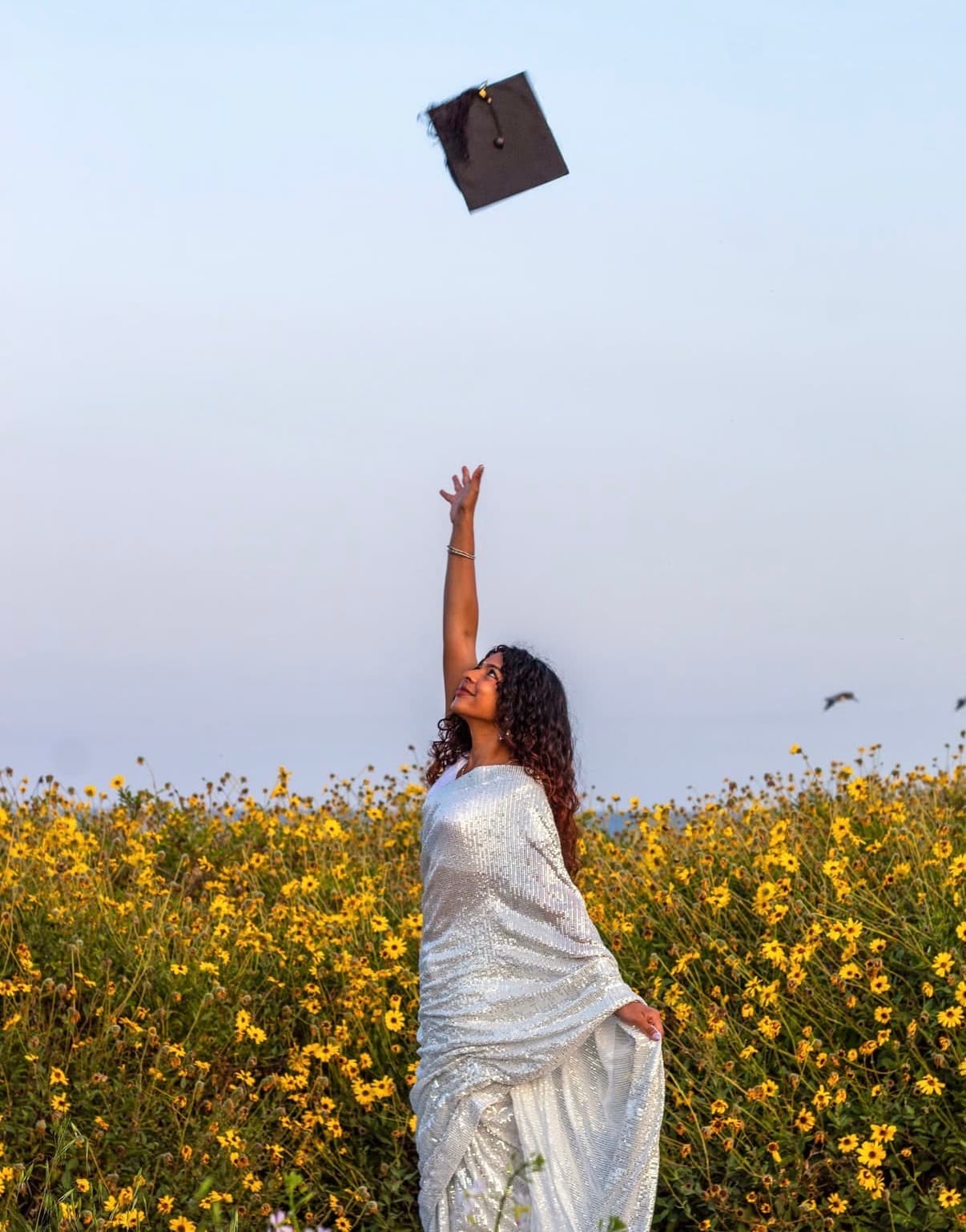 Graduate in a silver gown tossing her cap above a wildflower field at sunset.